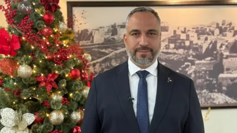 Bethlehem's mayor Maher Canawati stands next to a Christmas tree with red and gold decorations