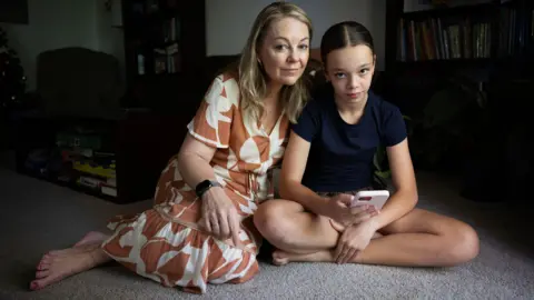 BBC/Jessica Hromas A  woman in a brown and white dress sits on a carpeted floor with her teenage daughter, who is holding a pink phone