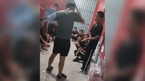 A still from mobile phone footage showing an instructor's back view as he stands with a rod raised, next to a student whose hand is outstretched. Other students are crouched and seated around the edge of the small room, which has red painted walls and a metal grille on the doorway. All the students' faces have been blurred.