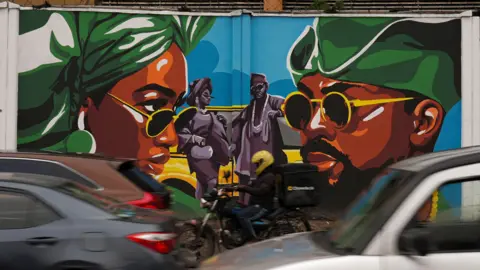 Sodiq Adelakun / REUTERS Cars and a motorbike drive past a mural of a man and a woman both wearing green headgear and sunglasses looking at each other, while a couple is dancing in between them.