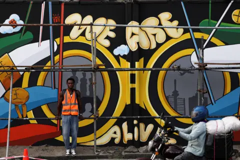 Sodiq Adelakun / REUTERS A man stands in front of a mural of a pair of binoculars outlined in yellow against a black background, along with the slogan: We Are Alive
