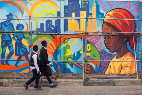 Toyin Adedokun / AFP via Getty Images Two men walk past a brightly coloured mural of a young woman painting the earth.