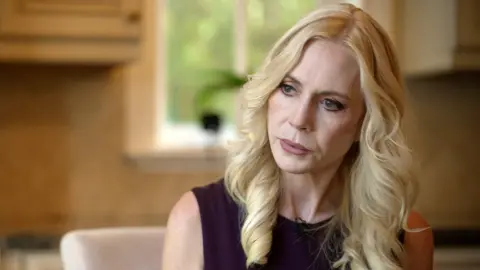 Sigrid McCawley, a woman with wavey blond hair and wearing a black dress, pictured in close-up in an office, looking to the left of the camera, with the background out of focus.