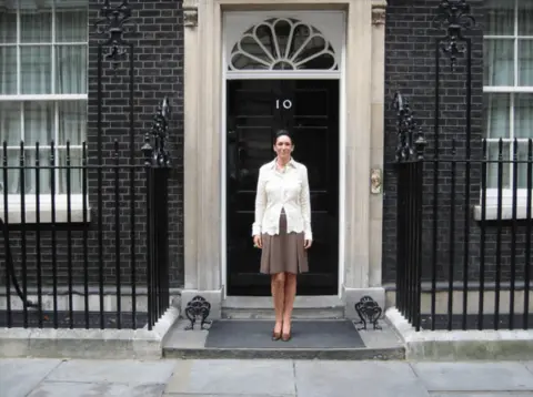 US Department of Justice Maxwell in white shirt and brown skirt and heels poses alone in front of the Downing St door