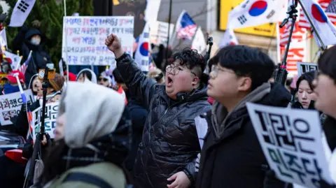 Getty Images A man in glasses and a black puffer jacket punches the air while yelling, surrounded by a crowd of people holding banners and South Korean flags