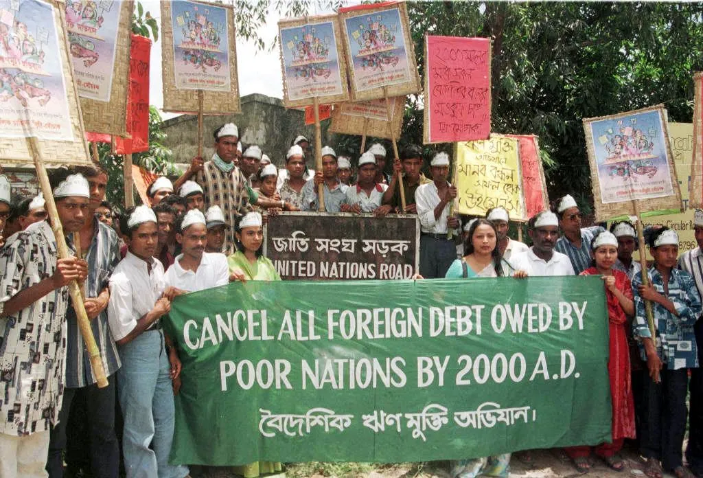 A group of protesters stand with a sign saying “Cancel All Foreign Debt Owed By Poor Nations By 2000 A.D.”