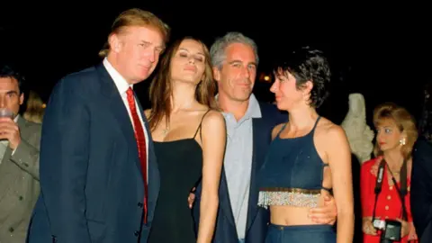 Getty Images A younger unsmiling Trump on the left in a suit with red tie, next to Melania in low-cut black dress with spaghetti straps, who has her eyes closed and is leaning her head toward Jeffrey Epstein, who looks out at the camera wearing a polo shirt and blazer and has his arm around the waist of Ghislaine Maxwell, who looks toward the group and is wearing a short denim halter top with beaded fringe