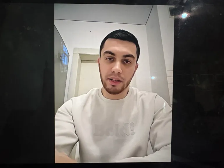 A photo of a computer screen with the image of Ilyas Abu Safiya on a video call. A clean-shaven young man with dark hair. Reflected in the computer screen is a streetlight because the journalists could only get enough internet to run an online interview by standing in the street, due to Israel's blockade of all services and goods in Gaza