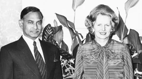 Getty Images A black and white photo from 1980 showing President Ziaur Rahman of Bangladesh wearing a suit and tie standing next to British Prime Minister Margaret Thatcher wearing a long-sleeved dress, standing in front of several leafy potted plants