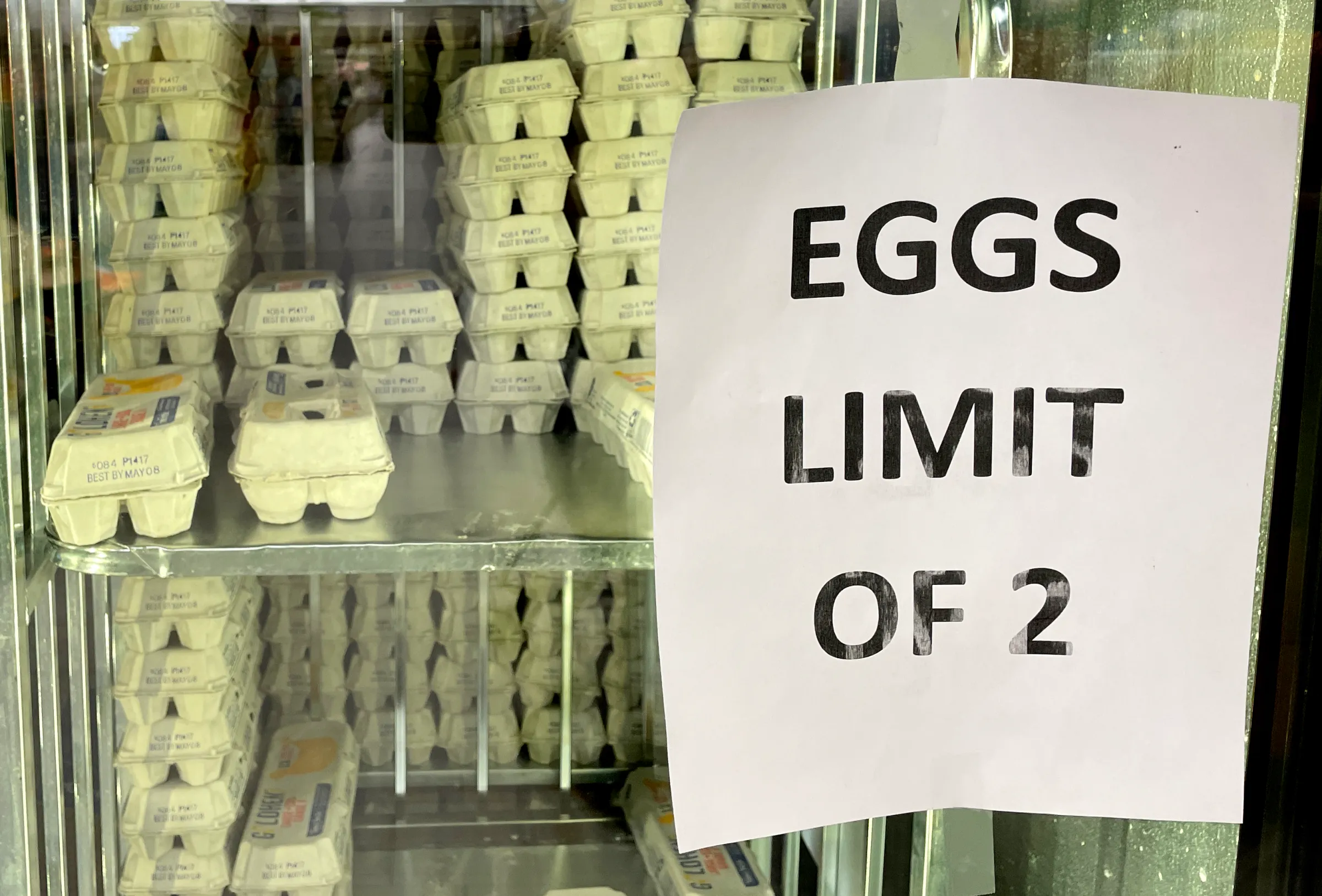 A grocery store cooler full of eggs, with a paper sign reading “Eggs Limit of 2.”
