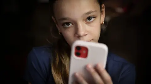 BBC/Jessica Hromas A teen wearing a navy shirt looks at the camera, her pink phone partially covering her face