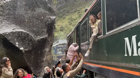Reuters A woman screams as she exits a crashed train through the window. Others beneath her try to help, reaching up towards her. There is a clear sense of panic on their faces. Behind them, a crashed train and a large cliff. 