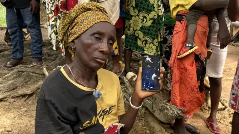 Andre Lombard / BBC A woman in a yellow, patterned headscarf and half-brown, half-yellow T-shirt is holding up the cracked screen of her smartphone.