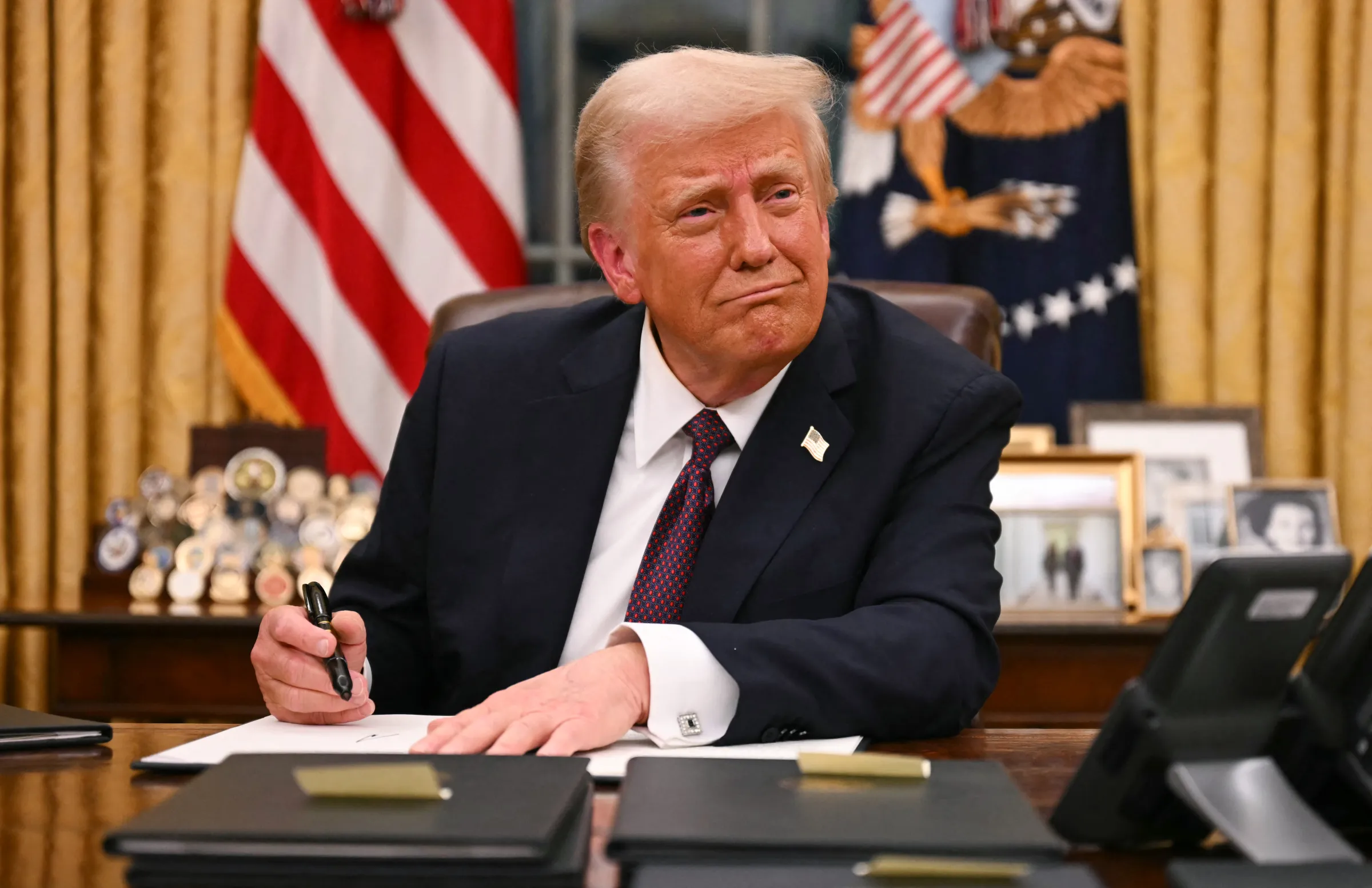 President Donald Trump sits at the Oval Office desk, pen in hand, smiling and signing papers in black folders.