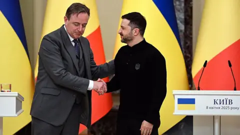 Ukrinform/NurPhoto Two men - one in a three-piece suit in grey, the other in black stand in front of colourful flags