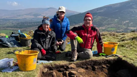 Antiquity PhD researcher Cherie Edwards (left) in black jacket and blue bandana,, her primary supervisor Dr Drik Brandherm in a red jacket and hat, and grey trousers, and research fellow Dr Linda Boutoille in a blue jacket, undertook an excavation at the site - they are kneeling beside a large hole with mountains in the background