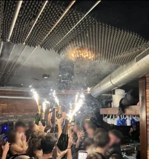 People hold sparklers attached to champagne bottles in the Swiss ski resort bar just before the fire started - with a small orange patch of fire seen on what looks like a foam ceiling above the sparklers.