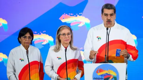 Gaby Oraa/Bloomberg via Getty Images Nicolas Maduro, right, speaks to members of the media, next to Cilia Flores, center, and Delcy Rodriguez, after casting a ballot during a referendum vote in Caracas, Venezuela, on Sunday, Dec. 3, 2023. They are wearing matching track suits with a rainbow printed on it. 