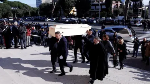 Reuters Pallbearers carry the coffin of 16-year-old Riccardo Minghetti up the steps of the Basilica of Saints Peter and Paul in Rome, with mourners all around, a hearse near a zebra-crossing, and other cars parked on the other side of the road.