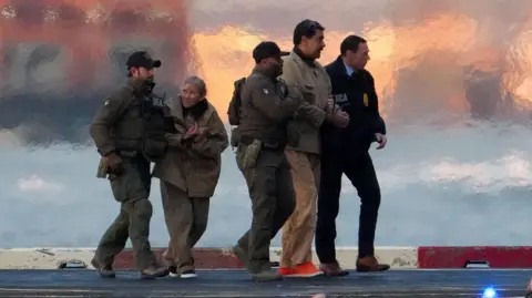 Reuters Three law enforcement officers escorting handcuffed Nicolás Maduro and his wife Cilia Flores on tarmac in New York City.