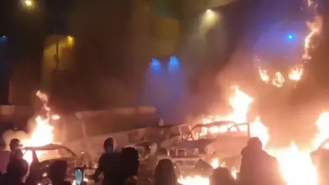 Eyewitness image / Reuters Protesters gathered in front of burning vehicles in Tehran at night in video released on 9 January 2026. Thick black smoke billows from the hulks of several burning cars with a large vehicle in the background. Around a dozen people are in the foreground. Lights from buildings are in the background.
