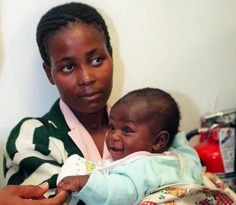 AFP via Getty Images A young mother is holding her smiling baby. The mum is wearing a stripy jacket and pink shirt and the baby is wearing a light blue top.