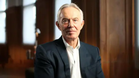 BBC/Monika Ghosh Tony Blair looks toward the camera. He is wearing a navy blue suit and open-collar white shirt.