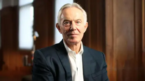 BBC/Monika Ghosh An image of former UK prime minister Tony Blair looking at the camera while dressed in a black suit and white shirt in a room with wooden walls. 