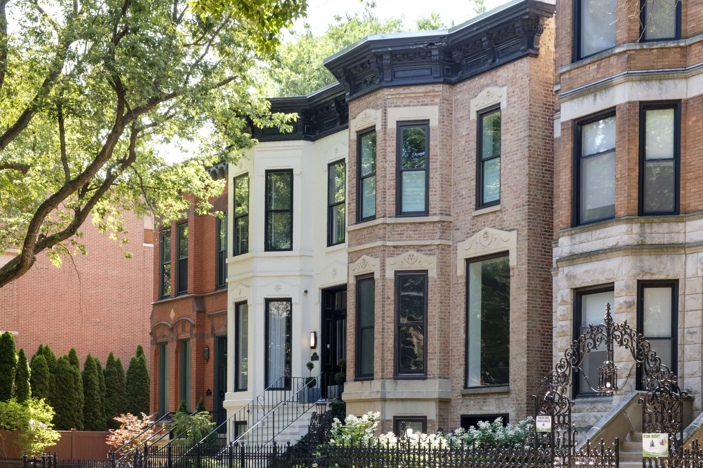 Tree-shaded row of historic brick and stone townhouses with bay windows, stoops, and decorative ironwork along a leafy residential street.