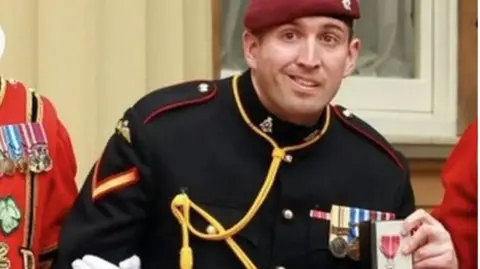 Getty An image of paratrooper Ben Parkinson from the chest up. He is in uniform, a black jacket with red trim on the epaulettes on each shoulder and with a gold rope trailed across his chest, and a burgundy beret on his head. He has medals pinned on his uniform and is holding an MBE. 