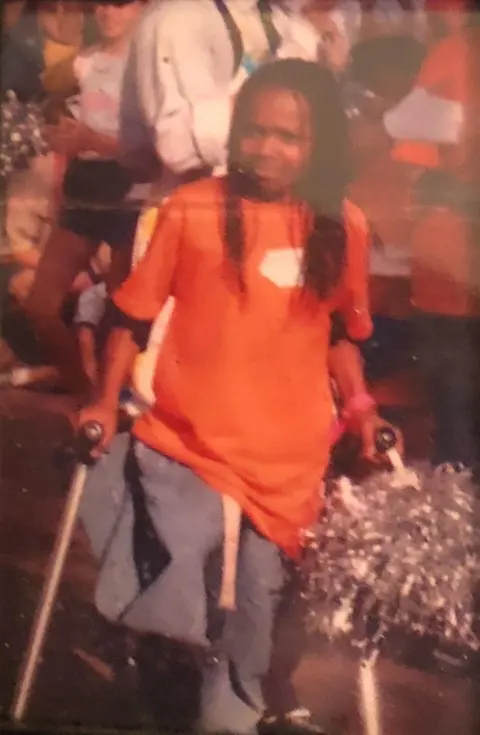 Abena Christine Jon'el Abena Christine Jon'el as young girl using crutches. She is wearing an orange T-shirt and the trousers on her right leg are folded up at the knee.