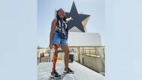 Abena Christine Jon'el Abena Christine Jon'el standing on a platform with a prosthetic right leg wrapped in kente cloth. She is pointing behind her at the top of Accra's Black Star monument.