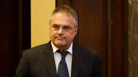 Reuters A man with a black jacket and tie and dark hair and glasses stares at a camera