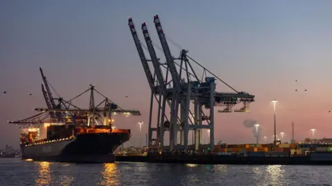 Bloomberg via Getty Images Cranes hover over a container ship with lights at dusk at the HHLA Container Terminal Tollerort (CTT) at the Port of Hamburg in Hamburg, Germany, on Monday, Feb. 3, 2025. 