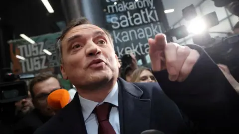 Reuters Zbigniew Ziobro wearing a blue coat, light blue shirt and burgundy tie, points with the raised index finger of his left hand 