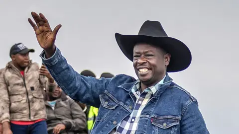 AFP/Getty Images Rigathi Gachagu in a cheque blue shirt, jean jacket and black cowboy hat waves at supporters in 2025.