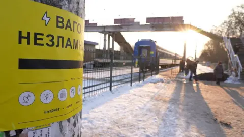BBC Yellow sign to left of picture says "Invincibility Train", while the train stands in the distance further to the right, on a very cold day with hard snow on the ground.