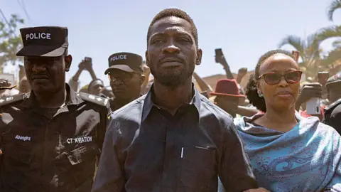 Getty Images Bobi Wine in a black shirt flanked by a police officer and his wife in a blue shawl on voting day.
