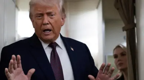 Reuters President Trump talks to reporters on Air Force One on Saturday. He is holding both hands up and Press Secretary Karoline Leavitt is watching on from behind.