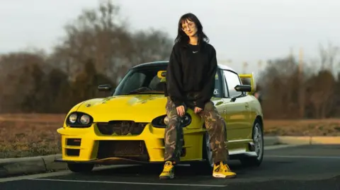 Jason Garcia Kei car enthusiast Nevi Bergeron sits on the bonnet of her mini sports car, a bright yellow modified Suzuki Cappuccino, in a car park next to a field.