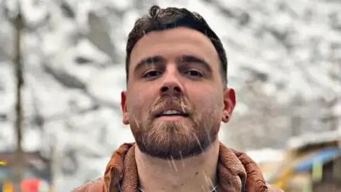Hengaw Organization for Human Rights/X A man with dark hair, a beard and a pierced ear looks at the camera as snow falls around him.