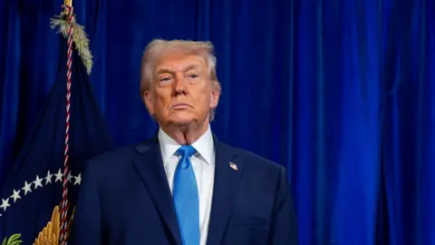 EPA US President Donald Trump, wearing a dark blue suit, white shirt, and light blue tie, looks ahead, his face stoic, at a news conference at the Mar-a-Lago Club in Palm Beach, Florida, USA. Behind him is a flag and blue curtains.