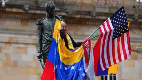 Getty Images A man wipes his tears while holding flags of Venezuela and the United States next to the statue of Simon Bolivar in Bogota, Colombia during a rally after Nicolás Maduro's capture.