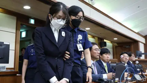 Getty Images A woman in a black suit, face mask and glasses is escorted into a court by a police officer who is holding her by the arm. Four men in suits sit at a table behind them