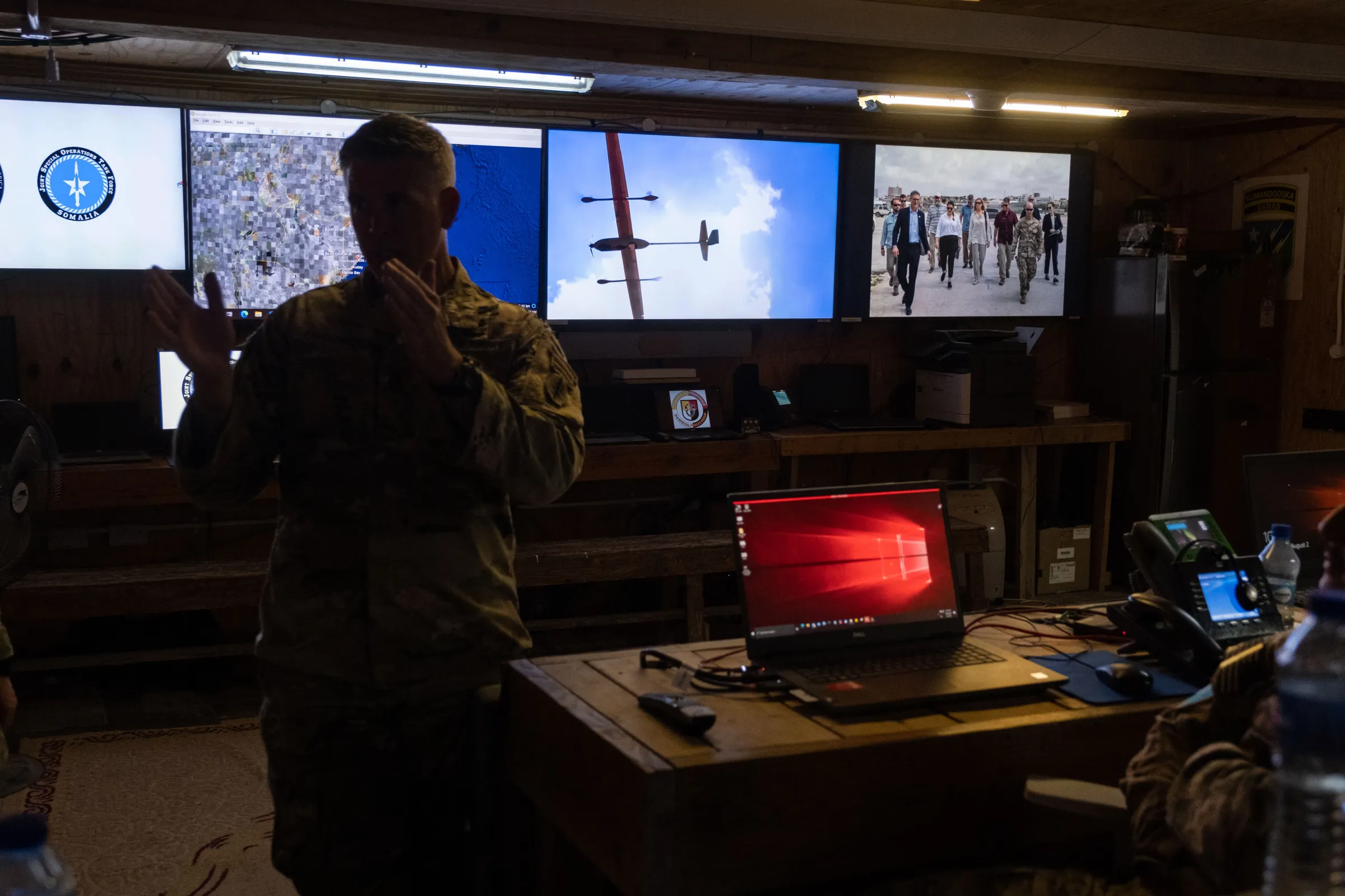 A man in camo fatigues stands in front of four screens.