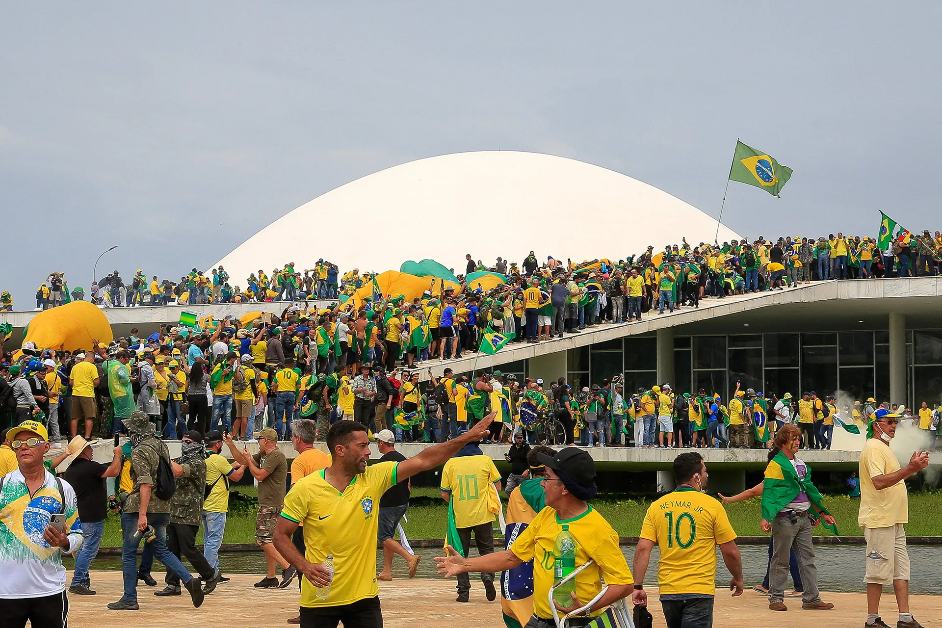 Large crowd of people dressed in yellow and green gather on and around a modern government building with a white dome, waving Brazilian flags