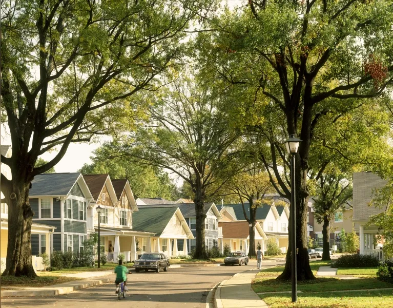 A quiet residential street lined with large, leafy trees and a row of pastel-painted, two-story houses with front porches. In the foreground, a child rides a bicycle along the curved road, while farther down the block an adult walks near the sidewalk; a few parked cars and a black lamppost sit beneath the tree canopy in warm late-afternoon light.
