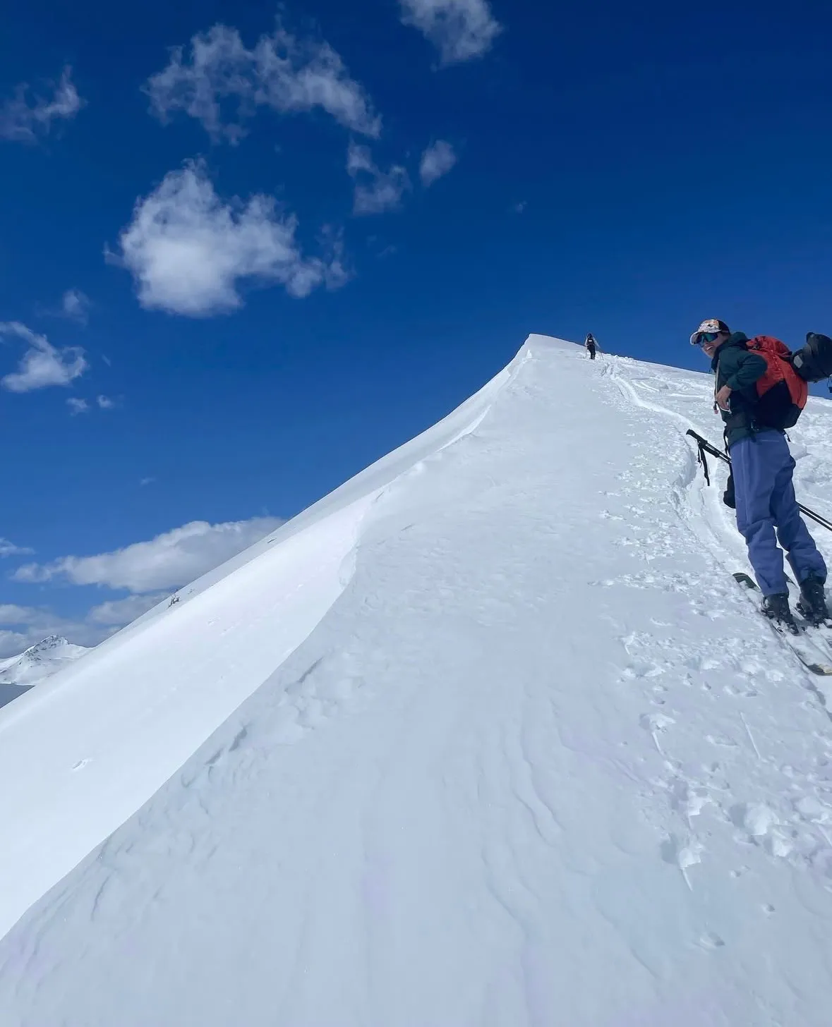 The sharp incline of a snow-covered peak with a person in the forground smiling back at the camera while on skis