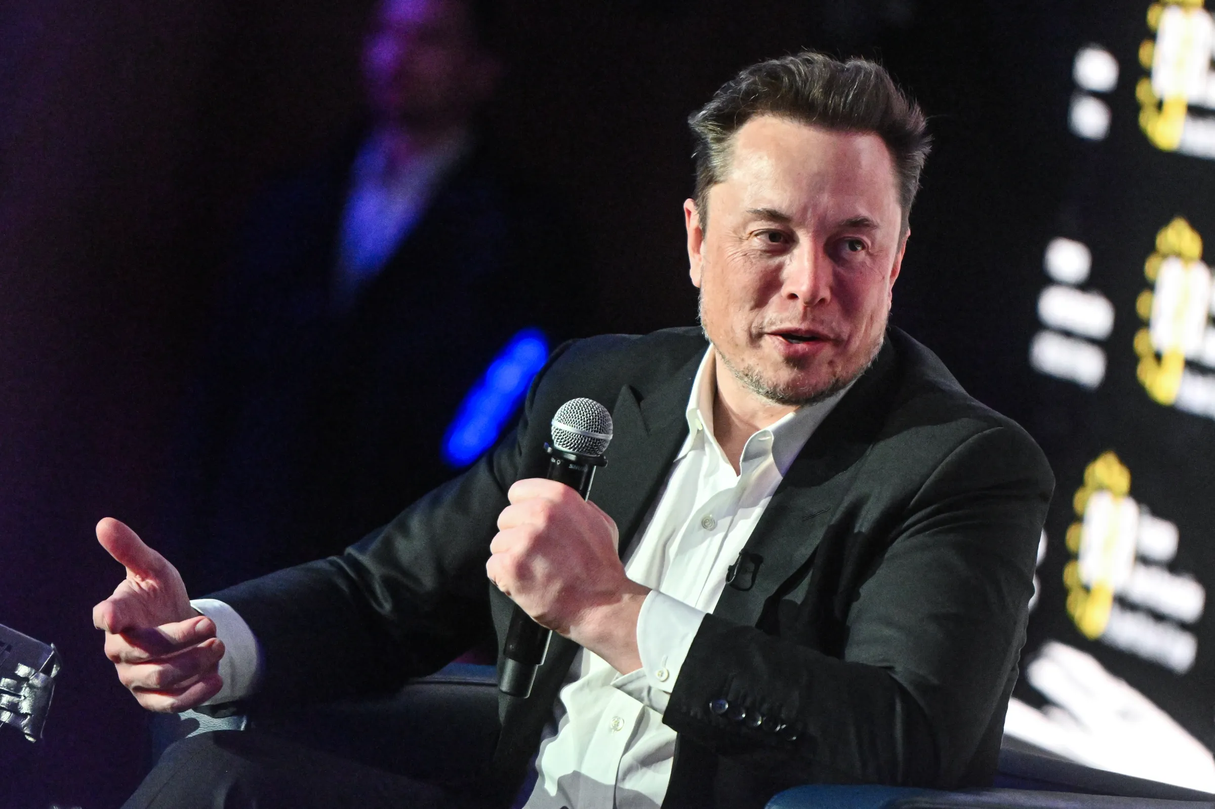 Elon Musk sitting and speaking, holding a microphone