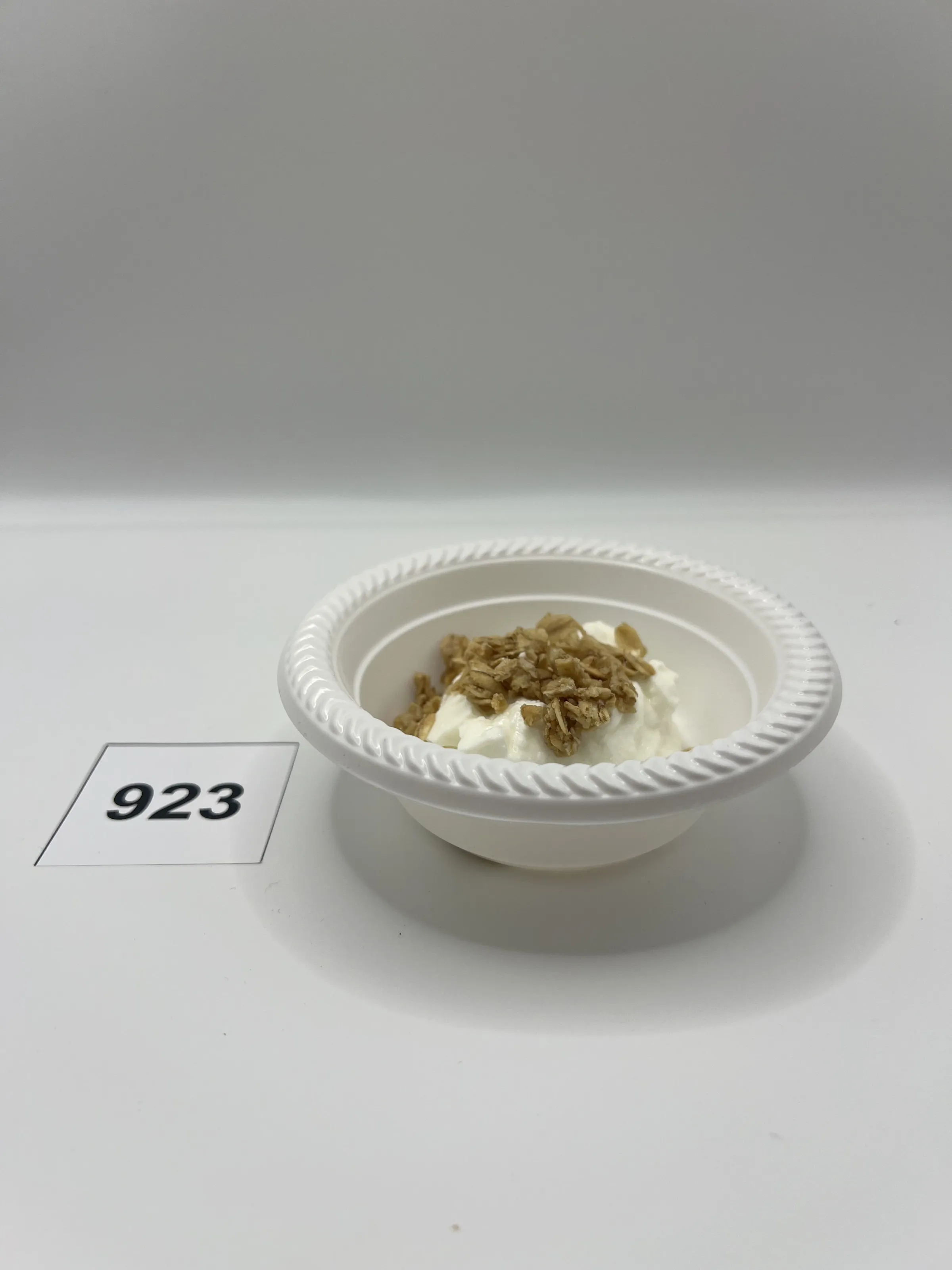 A small bowl of yogurt topped with granola. A piece of paper with the number 923 is next to the bowl.